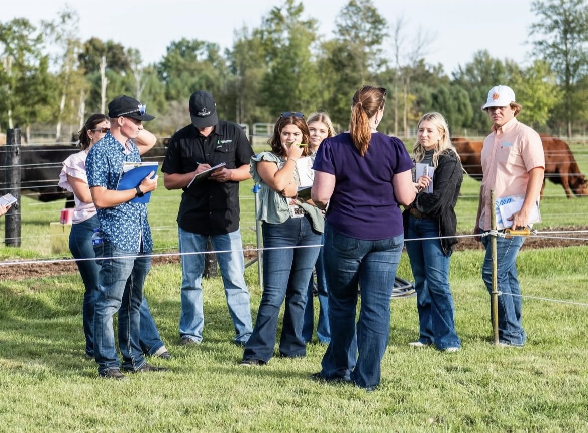 Livestock judging participants stand in a grassy outdoor pen, listening to instruction while holding notebooks